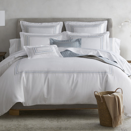 Mirabelle Collection by Home Treasures features white, sateen bedding with a blue-grey embroidered border detailing, styled with layered coordinating shams and pillows in soft neutral tones.